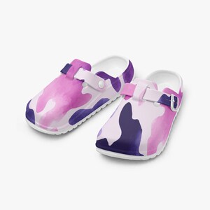 May include: A pair of clogs with a white base and a colorful pattern. The pattern features shades of pink, purple, and white in a camouflage design. The clogs have adjustable straps and a rounded toe.