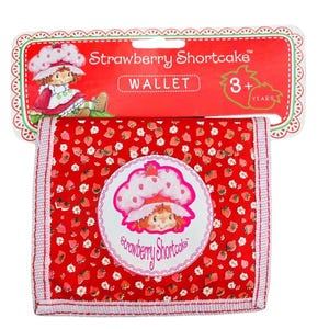 Pode incluir: Uma carteira vermelha da Strawberry Shortcake com um padrão de morangos. A carteira apresenta uma imagem circular da Strawberry Shortcake e o texto "Strawberry Shortcake". A embalagem diz "WALLET" e "3+ YEARS".