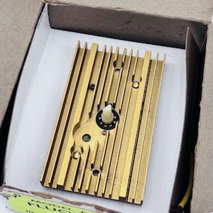 Puede incluir: Un disipador de calor rectangular dorado con aletas verticales y un mecanismo central. El dispositivo tiene pequeños agujeros y un componente de plástico blanco. Está dentro de una caja de cartón.