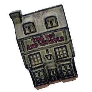 May include: A rectangular, vintage-style sign featuring the text "THE PIG AND WHISTLE" in red lettering. The sign depicts a building with multiple windows and a chimney, with a weathered, aged appearance.