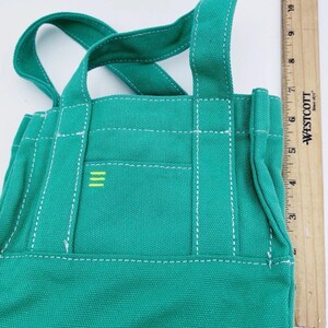 May include: A small, teal canvas tote bag with white stitching and two handles. The bag features a small front pocket with three horizontal yellow stripes. The bag is approximately 20 cm tall and 18 cm wide.