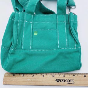 May include: A teal canvas tote bag with white stitching and a small yellow stripe design. The bag has two short handles and a long adjustable strap. A wooden ruler is placed next to the bag, measuring up to 20 cm.