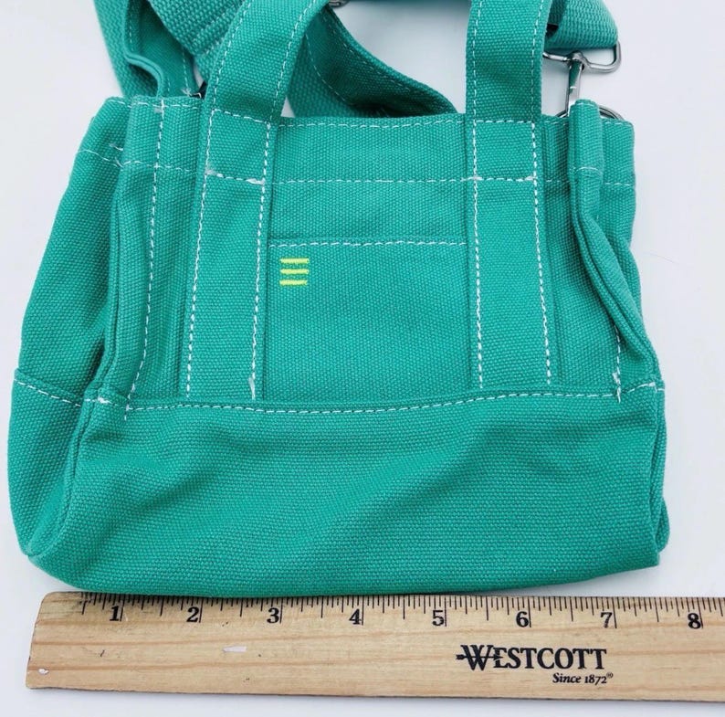 May include: A teal canvas tote bag with white stitching and a small yellow stripe design. The bag has two short handles and a long adjustable strap. A wooden ruler is placed next to the bag, measuring up to 20 cm.