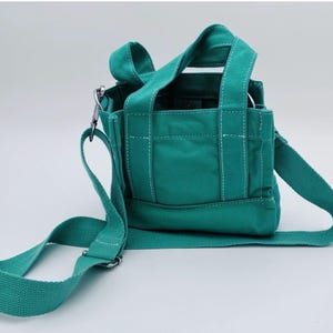 May include: A teal canvas tote bag with a shoulder strap and top handles. The bag features white stitching details and silver-tone metal hardware. The bag is designed with a rectangular shape and a flat bottom.