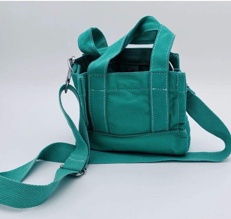 May include: A teal canvas tote bag with a shoulder strap and top handles. The bag features white stitching details and silver-tone metal hardware. The bag is designed with a rectangular shape and a flat bottom.