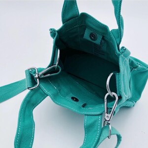 May include: Teal canvas tote bag with a detachable shoulder strap and two short handles. The bag features silver-tone metal hardware, including clasps and rings. The interior is visible, and the bag appears to be empty.