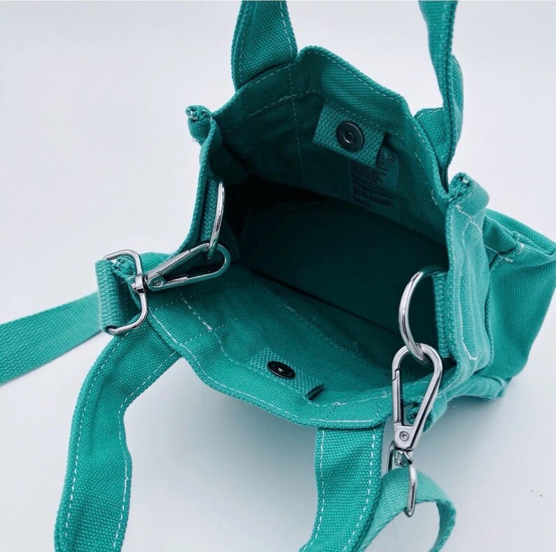 May include: Teal canvas tote bag with a detachable shoulder strap and two short handles. The bag features silver-tone metal hardware, including clasps and rings. The interior is visible, and the bag appears to be empty.