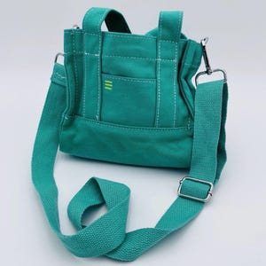 May include: Teal canvas tote bag with a detachable shoulder strap and top handles. The bag features a small front pocket with three vertical yellow stripes and white stitching details. The bag has a silver-coloured metal clasp and buckle.