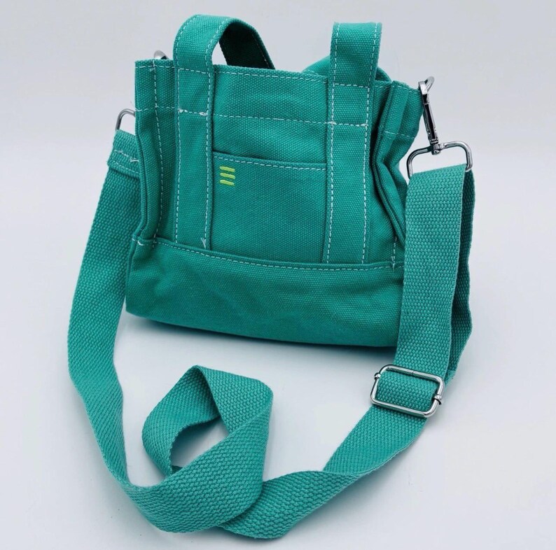 May include: Teal canvas tote bag with a detachable shoulder strap and top handles. The bag features a small front pocket with three vertical yellow stripes and white stitching details. The bag has a silver-coloured metal clasp and buckle.
