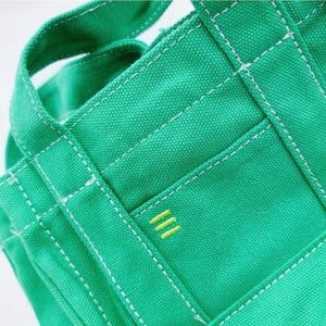 May include: Close-up of a green canvas tote bag with white stitching and a small pocket. The pocket features three horizontal yellow lines. The bag has sturdy handles and a structured design, suitable for carrying various items.