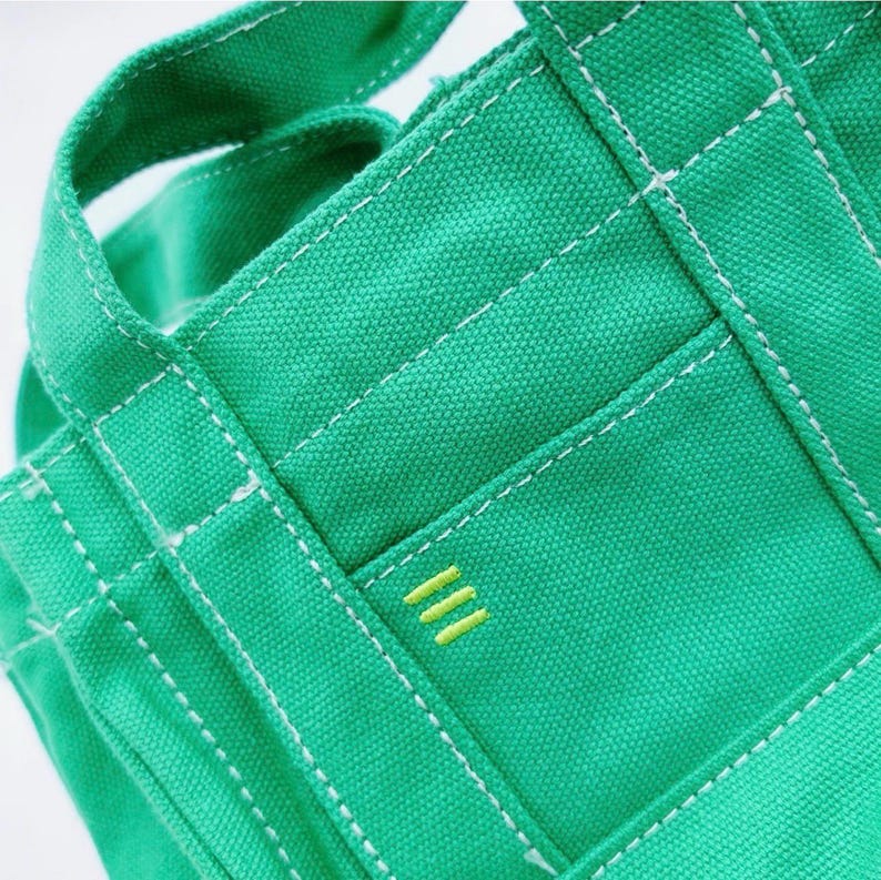 May include: Close-up of a green canvas tote bag with white stitching and a small pocket. The pocket features three horizontal yellow lines. The bag has sturdy handles and a structured design, suitable for carrying various items.
