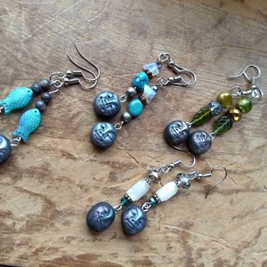 May include: Four pairs of dangle earrings with silver hooks. Each pair features a different design with various colored beads and charms. The first pair has blue fish charms and blue and silver beads. The second pair has turquoise and silver beads and a crescent moon charm. The third pair has green leaves and green and gold beads. The fourth pair has white and clear beads and a crescent moon charm.