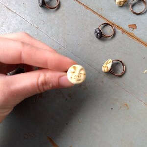 Puede incluir: Anillos hechos a mano con dise&ntilde;os &uacute;nicos. Los anillos presentan bandas de cobre y varios engastes de piedra o cer&aacute;mica, incluyendo una cara de luna color crema y piedras texturizadas de color gris oscuro. Se muestran sobre una superficie gris desgastada.