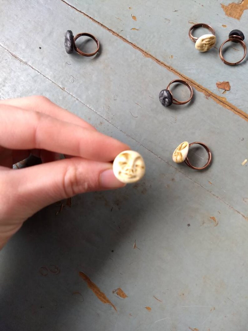 Puede incluir: Anillos hechos a mano con dise&ntilde;os &uacute;nicos. Los anillos presentan bandas de cobre y varios engastes de piedra o cer&aacute;mica, incluyendo una cara de luna color crema y piedras texturizadas de color gris oscuro. Se muestran sobre una superficie gris desgastada.