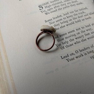 Puede incluir: Un anillo de cobre hecho a mano con una piedra de color claro. El anillo est&aacute; colocado sobre una p&aacute;gina de libro con el t&iacute;tulo "Leaders" visible. La banda del anillo tiene un dise&ntilde;o simple y circular.