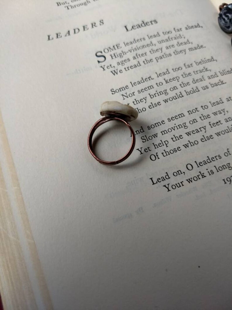 Puede incluir: Un anillo de cobre hecho a mano con una piedra de color claro. El anillo est&aacute; colocado sobre una p&aacute;gina de libro con el t&iacute;tulo "Leaders" visible. La banda del anillo tiene un dise&ntilde;o simple y circular.