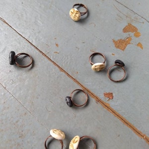 Puede incluir: Una colecci&oacute;n de anillos hechos a mano con bandas de color cobre. Algunos anillos presentan piedras circulares texturizadas de color blanquecino, mientras que otros tienen piedras oblongas de color gris oscuro. Los anillos est&aacute;n colocados sobre una superficie de madera gris envejecida.