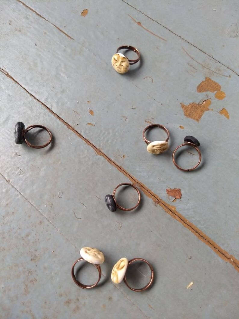Puede incluir: Una colecci&oacute;n de anillos hechos a mano con bandas de color cobre. Algunos anillos presentan piedras circulares texturizadas de color blanquecino, mientras que otros tienen piedras oblongas de color gris oscuro. Los anillos est&aacute;n colocados sobre una superficie de madera gris envejecida.