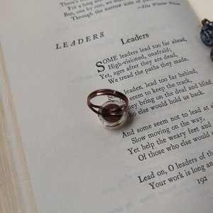 Puede incluir: Un anillo de tono cobrizo con un dise&ntilde;o circular impreso se encuentra sobre las p&aacute;ginas de un libro abierto. El libro muestra el poema "Leaders" de Ella Wheeler Wilcox. El dise&ntilde;o del anillo es visible.