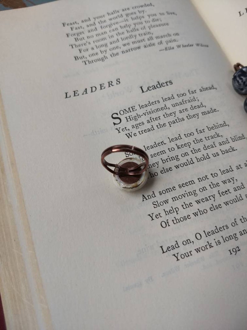 Puede incluir: Un anillo de tono cobrizo con un dise&ntilde;o circular impreso se encuentra sobre las p&aacute;ginas de un libro abierto. El libro muestra el poema "Leaders" de Ella Wheeler Wilcox. El dise&ntilde;o del anillo es visible.