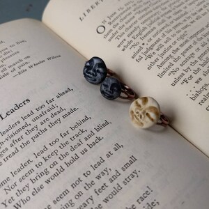 Puede incluir: Tres anillos con dise&ntilde;os de caras &uacute;nicos descansan sobre un libro abierto. Dos anillos presentan caras de color gris oscuro, mientras que el tercero tiene una cara de color crema. Los anillos tienen bandas de color cobre. El libro muestra texto sobre libertad y liderazgo.