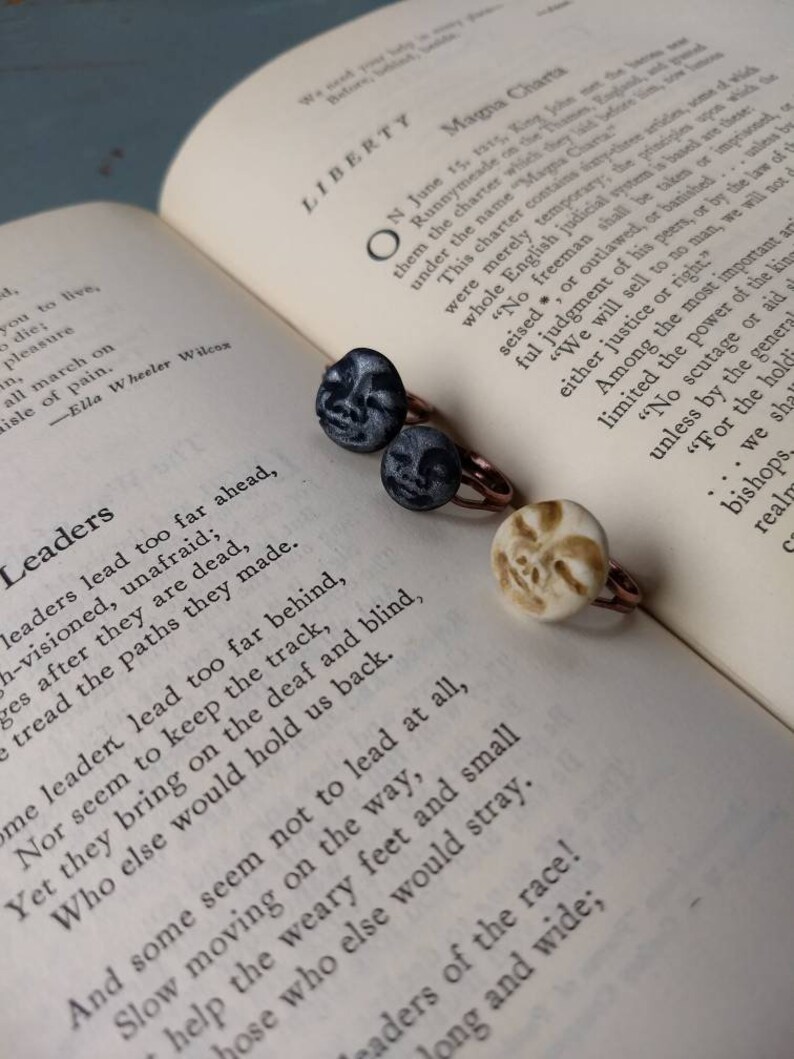 Puede incluir: Tres anillos con dise&ntilde;os de caras &uacute;nicos descansan sobre un libro abierto. Dos anillos presentan caras de color gris oscuro, mientras que el tercero tiene una cara de color crema. Los anillos tienen bandas de color cobre. El libro muestra texto sobre libertad y liderazgo.