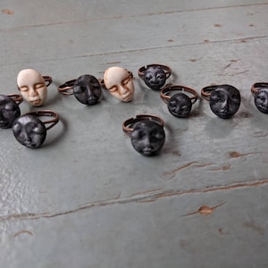May include: A collection of ten rings featuring faces, some with closed eyes and some with open eyes. The rings are made of copper and have a dark patina. The faces are made of white and black clay.