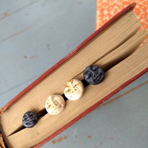 Puede incluir: Un libro antiguo abierto, con lomo rojo, contiene cuatro peque&ntilde;os botones redondos decorativos. Los botones son de color crema y gris oscuro, cada uno con un dise&ntilde;o de rostro. Las p&aacute;ginas del libro est&aacute;n envejecidas y otro libro es visible en el fondo.