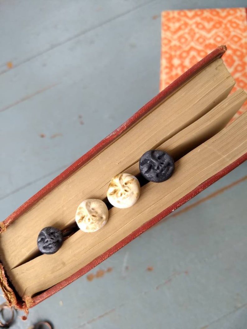 Puede incluir: Un libro antiguo abierto, con lomo rojo, contiene cuatro peque&ntilde;os botones redondos decorativos. Los botones son de color crema y gris oscuro, cada uno con un dise&ntilde;o de rostro. Las p&aacute;ginas del libro est&aacute;n envejecidas y otro libro es visible en el fondo.