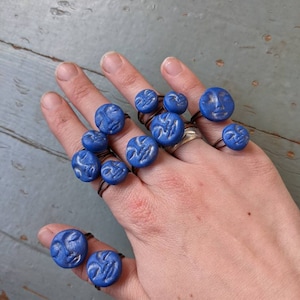 May include: A hand displays several blue rings with carved faces. The rings have a dark band and a round, textured blue face. The rings vary in size and are worn on multiple fingers.