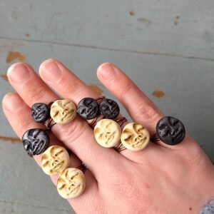 Puede incluir: Una mano muestra varios anillos con caras circulares en negro y crema. Los anillos tienen una banda de color cobre. Las caras tienen detalles tallados, y el fondo es una superficie de madera azul claro.