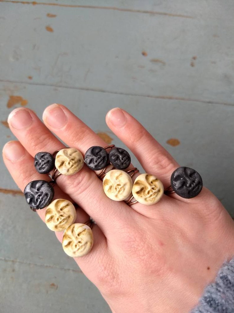 Puede incluir: Una mano muestra varios anillos con caras circulares en negro y crema. Los anillos tienen una banda de color cobre. Las caras tienen detalles tallados, y el fondo es una superficie de madera azul claro.