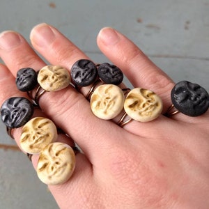May include: A hand displaying several rings with circular, face-shaped pendants. The rings have copper-colored bands. The pendants are in shades of black and cream, with carved facial features. The background is a light blue wooden surface.