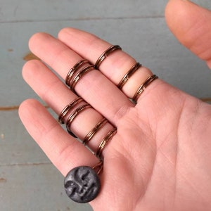 Puede incluir: Anillos de alambre color cobre que se usan en los dedos, conectados por una cadena a un colgante circular gris oscuro. El colgante tiene un dise&ntilde;o de rostro tallado. Los anillos se usan en varios dedos.