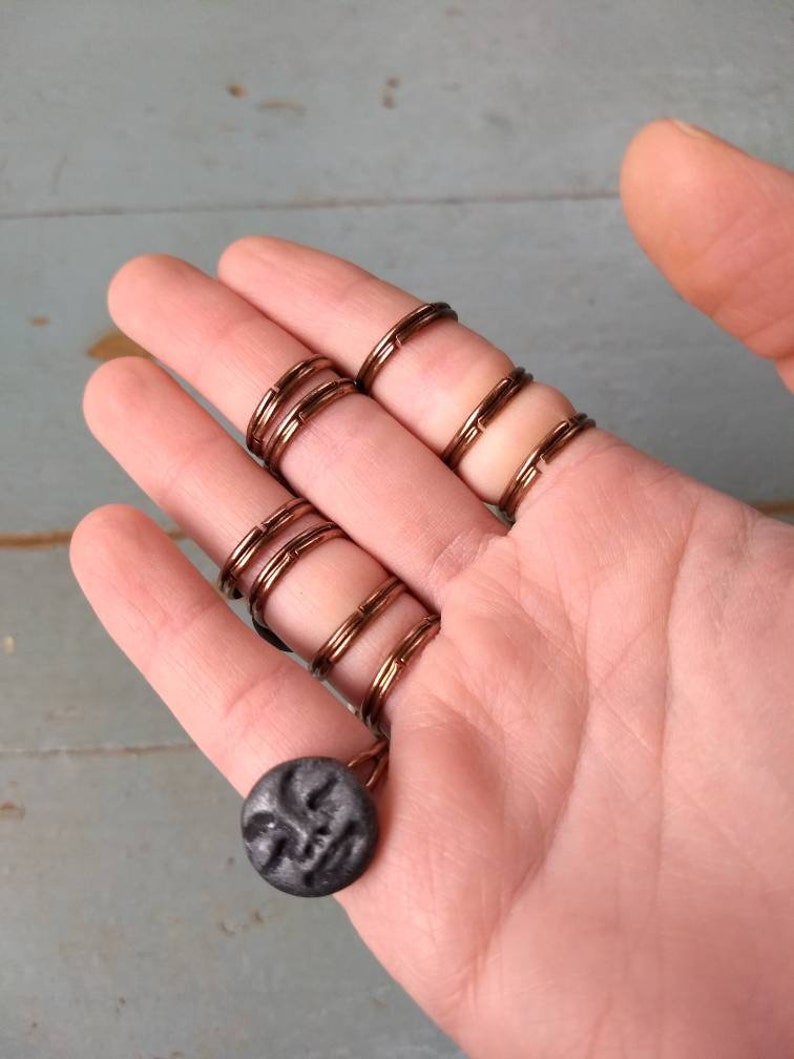 Puede incluir: Anillos de alambre color cobre que se usan en los dedos, conectados por una cadena a un colgante circular gris oscuro. El colgante tiene un dise&ntilde;o de rostro tallado. Los anillos se usan en varios dedos.