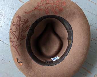 May include: A brown felt hat with a brown leather band and an embroidered oak leaf design in orange. The hat has a label inside that reads "Woolrich".