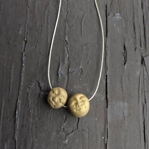 May include: A gold necklace with two round pendants featuring a face design. The necklace is on a wooden surface.
