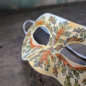May include: A cream-colored masquerade mask with a leaf design in orange, green, and black. The mask has a white string and eye openings. The leaves are detailed with black outlines and the mask is on a dark brown surface.
