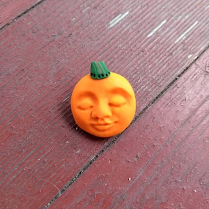May include: A small orange pumpkin with a green stem and a smiling face. The pumpkin is made of clay and has a smooth, matte finish.