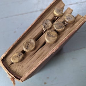 May include: A vintage book with six brown leaf-shaped bookmarks. The bookmarks are made of clay and have a rustic, earthy look.