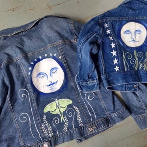 May include: Two denim jackets with hand-painted designs. The larger jacket features a white moon with a face, a green moth, and white swirls. The smaller jacket has a white moon with a face, white stars, and green swirls.