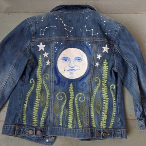 May include: A blue denim jacket with a hand-painted design featuring a white moon with a face, green ferns, and white stars on the back.
