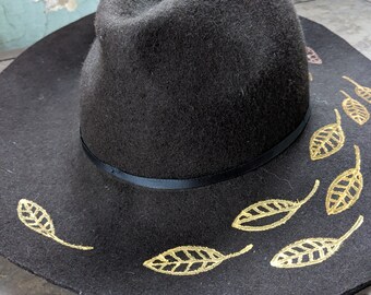 May include: A black felt fedora hat with a wide brim and gold leaf accents.