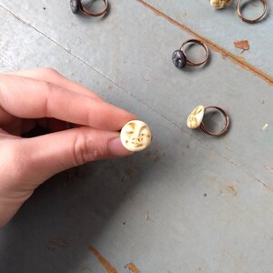 Puede incluir: Anillos hechos a mano con bandas de cobre y peque&ntilde;as caras esculpidas. Las caras son de color crema y gris oscuro. Una mano sostiene un anillo, mostrando los detalles. Los anillos est&aacute;n sobre una superficie azul claro desgastada.