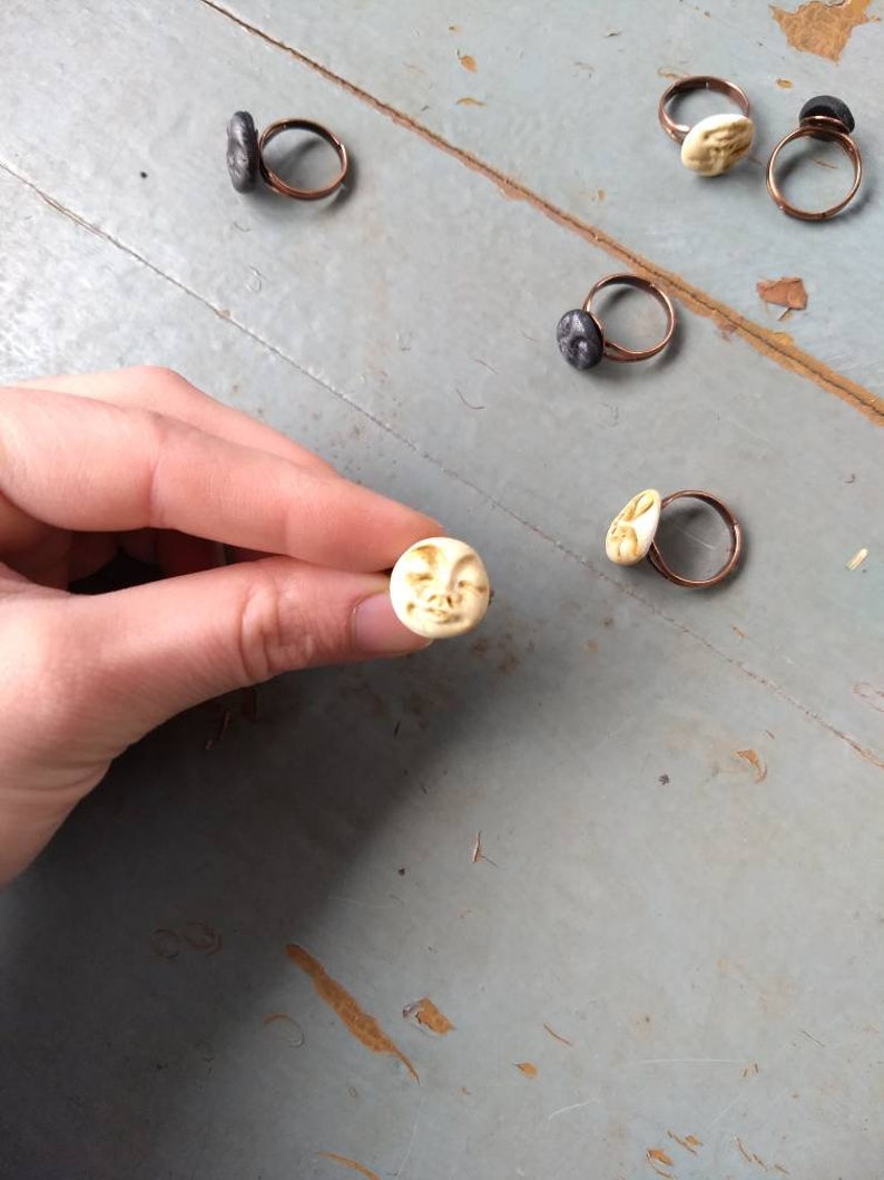 Puede incluir: Anillos hechos a mano con bandas de cobre y peque&ntilde;as caras esculpidas. Las caras son de color crema y gris oscuro. Una mano sostiene un anillo, mostrando los detalles. Los anillos est&aacute;n sobre una superficie azul claro desgastada.