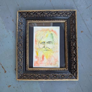 May include: A framed watercolor portrait of a man with a long beard and hair, rendered in shades of yellow, orange, and green. The artwork is set against a black mat and framed in a black and gold ornate frame.