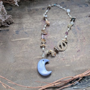 May include: A silver chain necklace with a blue crescent moon pendant and various brown, white, and iridescent beads. The necklace is on a rustic wooden surface.