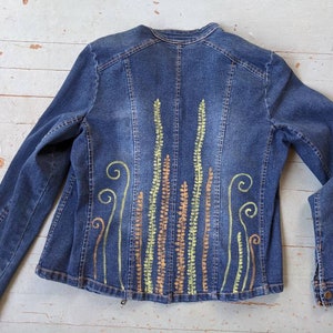 May include: A blue denim jacket with a painted design on the back. The design features green, orange, and brown lines and swirls.