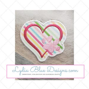 May include: Embroidered patch featuring a pink paw print within a pink and white striped heart. The heart is outlined in pink and white. The text "Lydia Blue Designs.com" is below the patch. Embroidery, sublimation and handmade crafts.