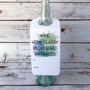 May include: A clear glass wine bottle with a white gift tag. The tag has the text "WINE. HOW CLASSY PEOPLE GET SHIT-FACED." with watercolor accents. The tag also has spaces for "TO:" and "FROM:"